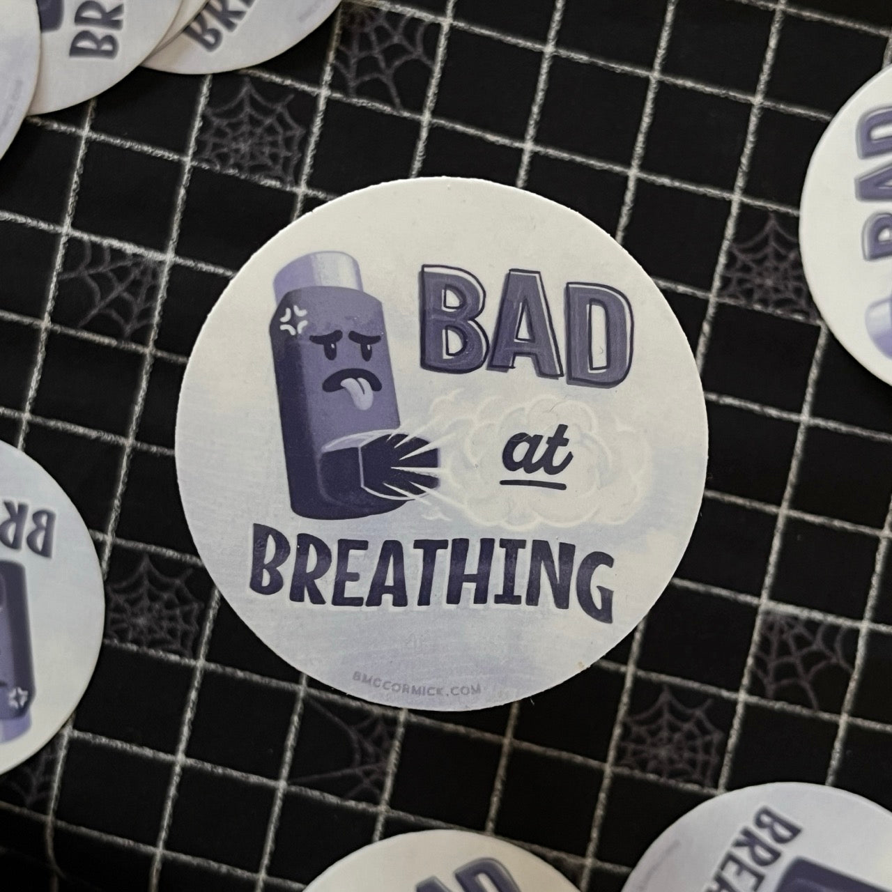 Wheezing inhaler on round sticker with "Bad at Breathing". Black checker background